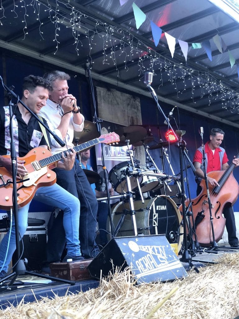 Playing band on stage with musical instruments under string lights and colourful bunting, in a covered outdoor setting.