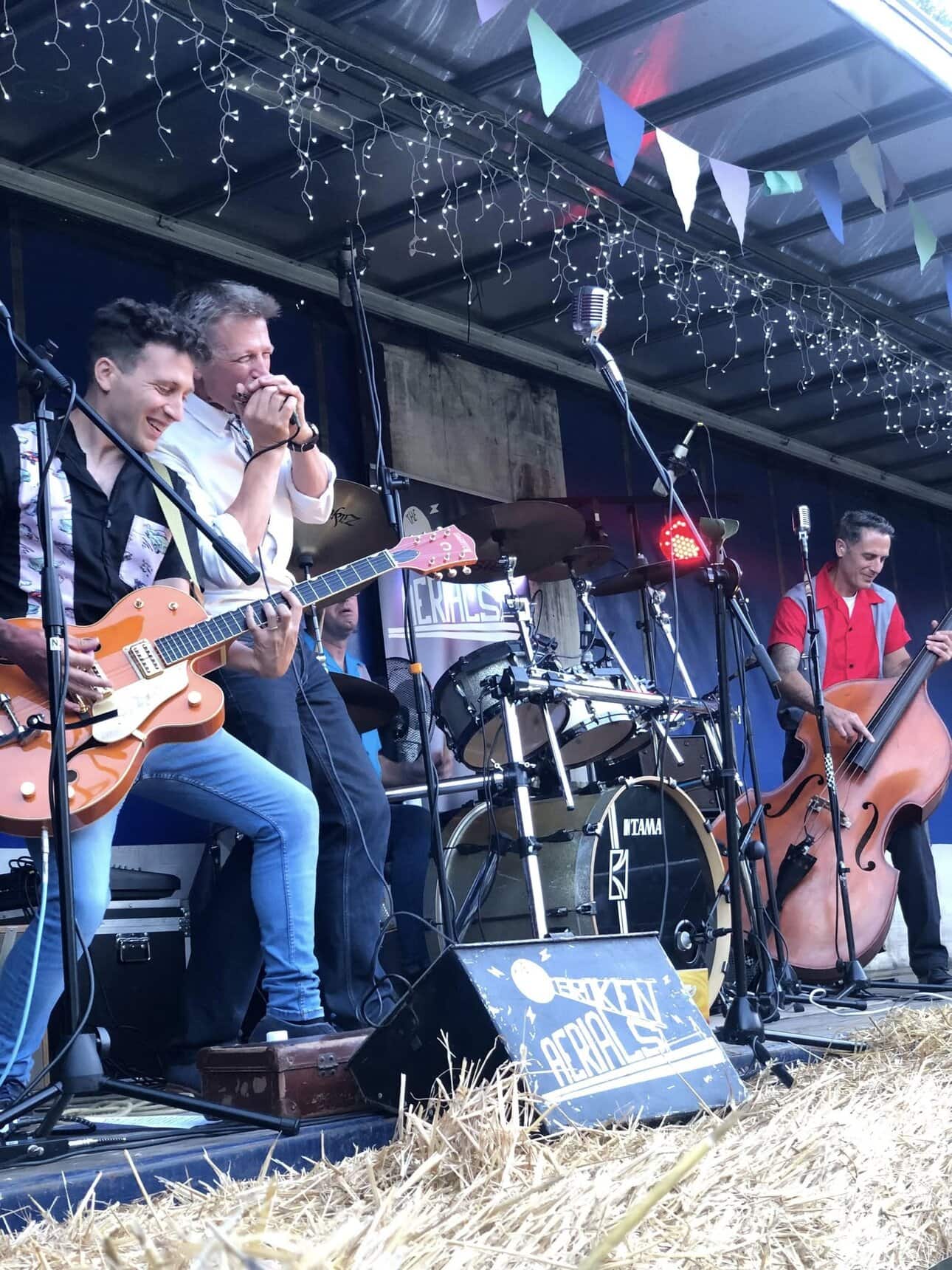 Playing band on stage with musical instruments under string lights and colourful bunting, in a covered outdoor setting.