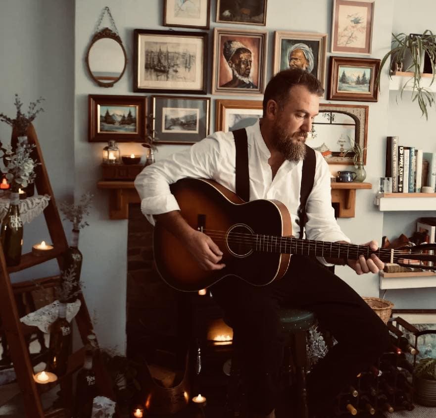 A man playing acoustic guitar in a cosy indoor setting with artwork on the wall.
