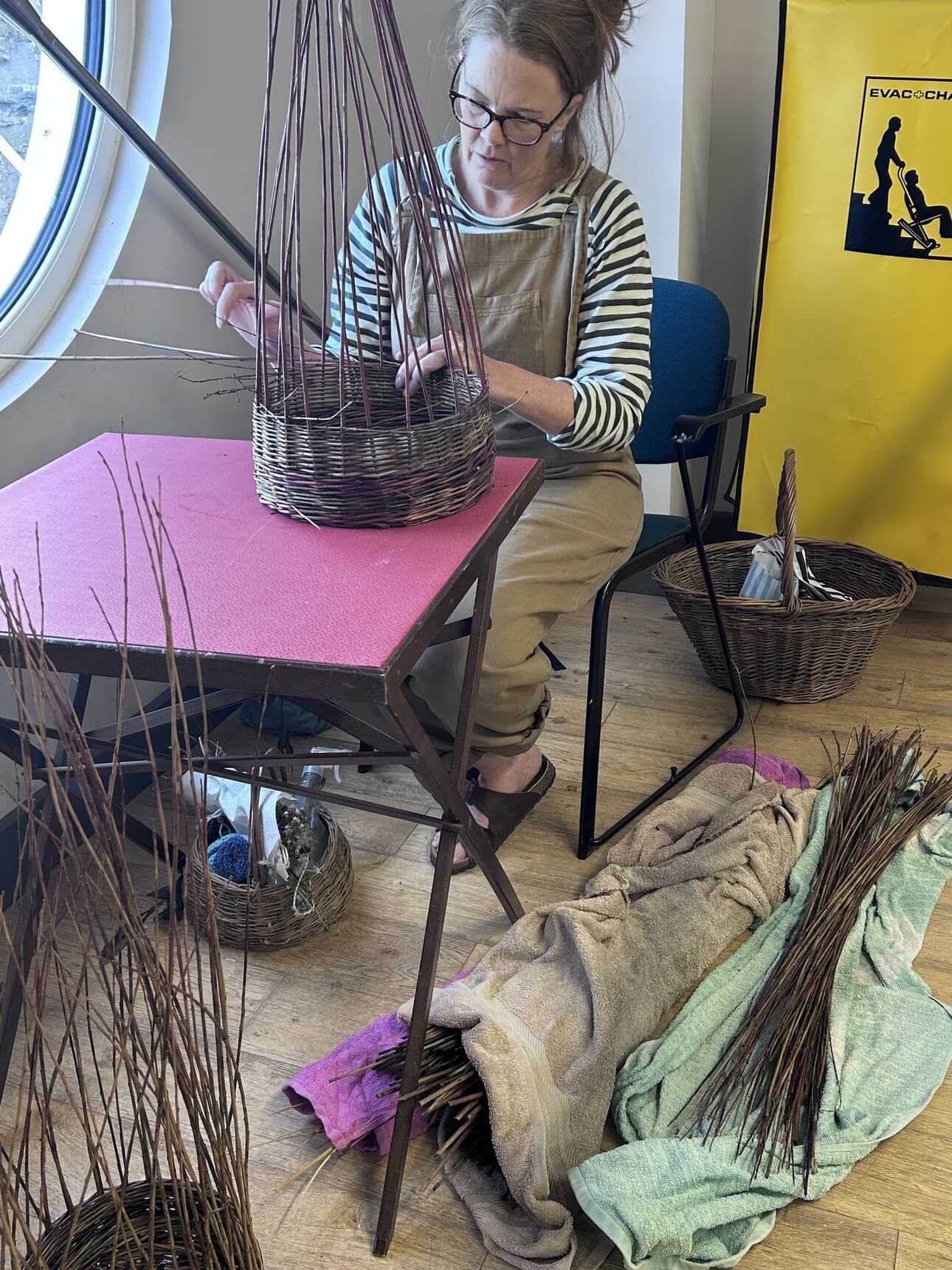 Woman weaving a basket with willow branches indoors, using a pink work mat and surrounded by baskets and towels.