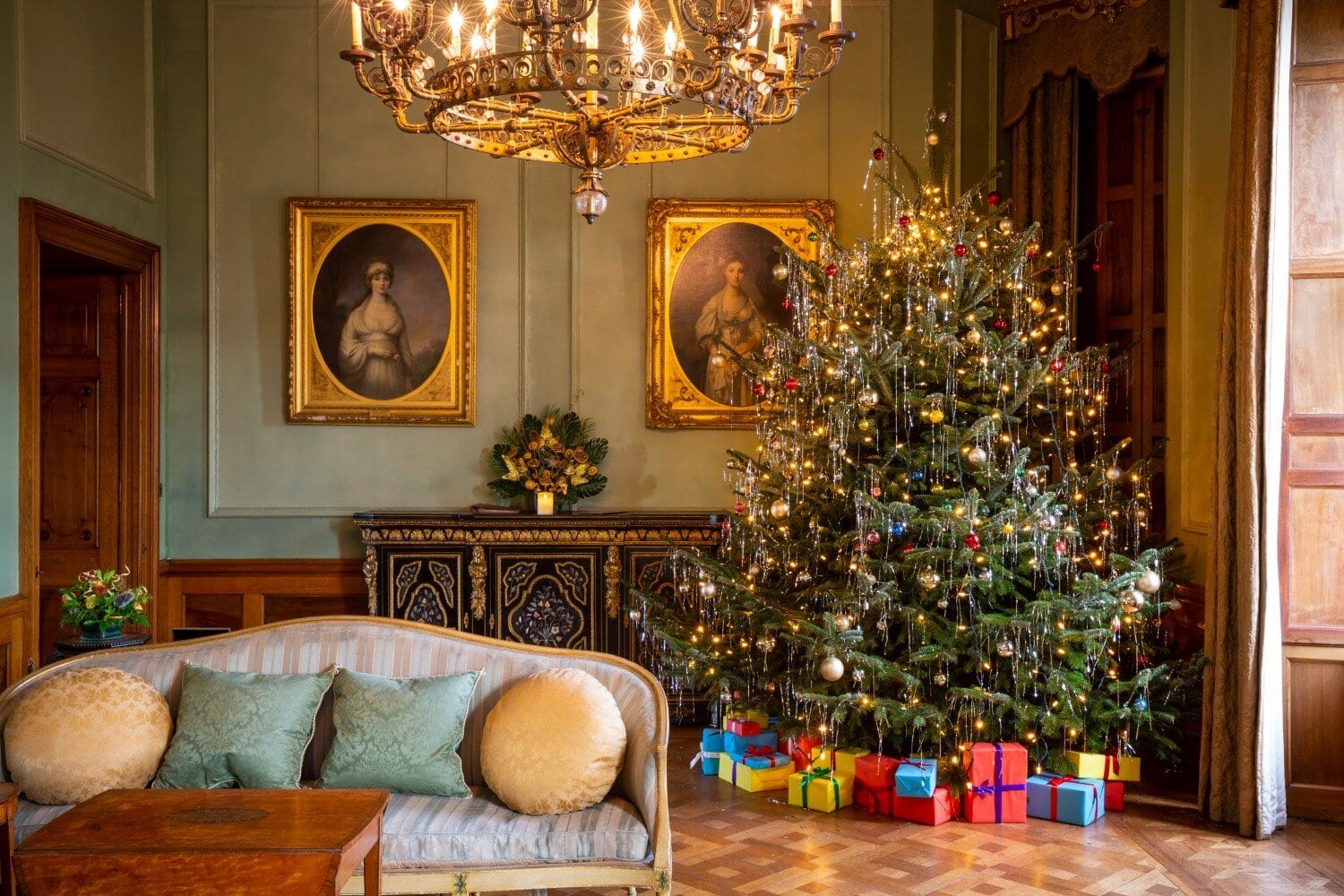 A beautifully decorated indoor Christmas scene with a large tree, presents, and vintage portraits in a historic hotel.