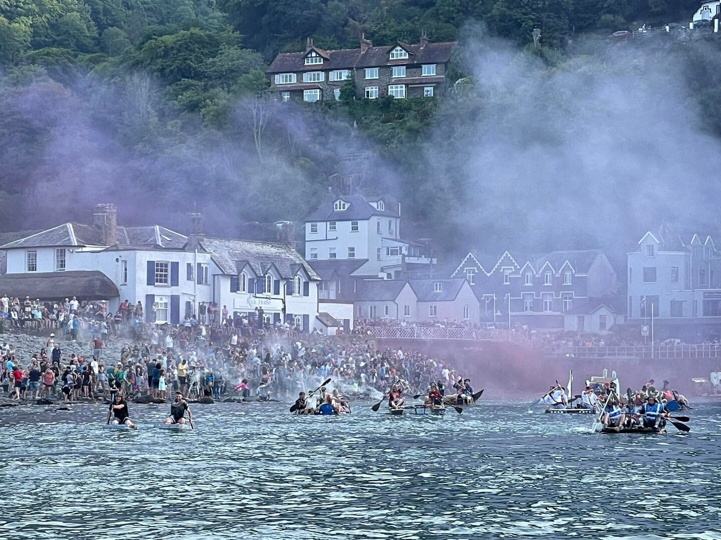 A bustling harbour scene in Lynmouth with boats, visitors, and charming hillside houses amid mist and greenery.