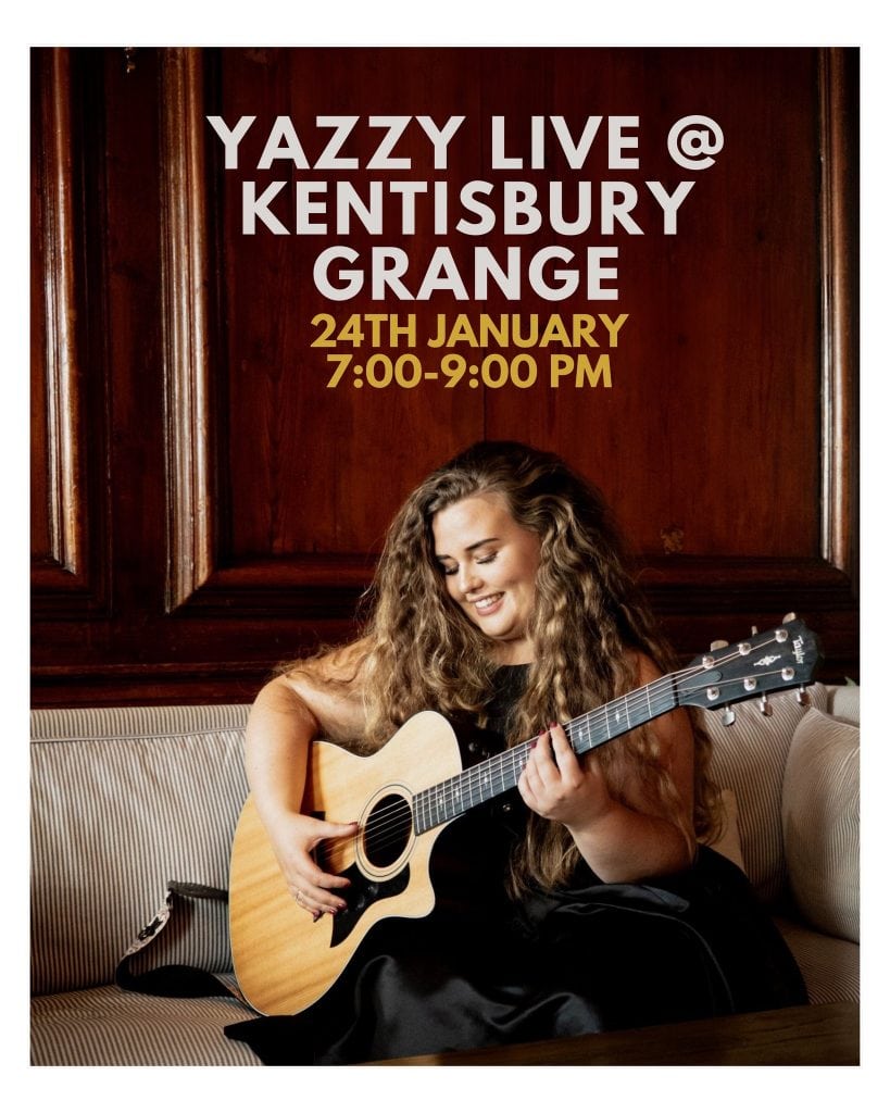 Young woman playing acoustic guitar, smiling, sitting on a sofa in a wood-paneled room.