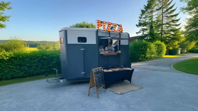 A mobile pizza stand with a "PIZZA" sign serving food outdoors in a lush park.