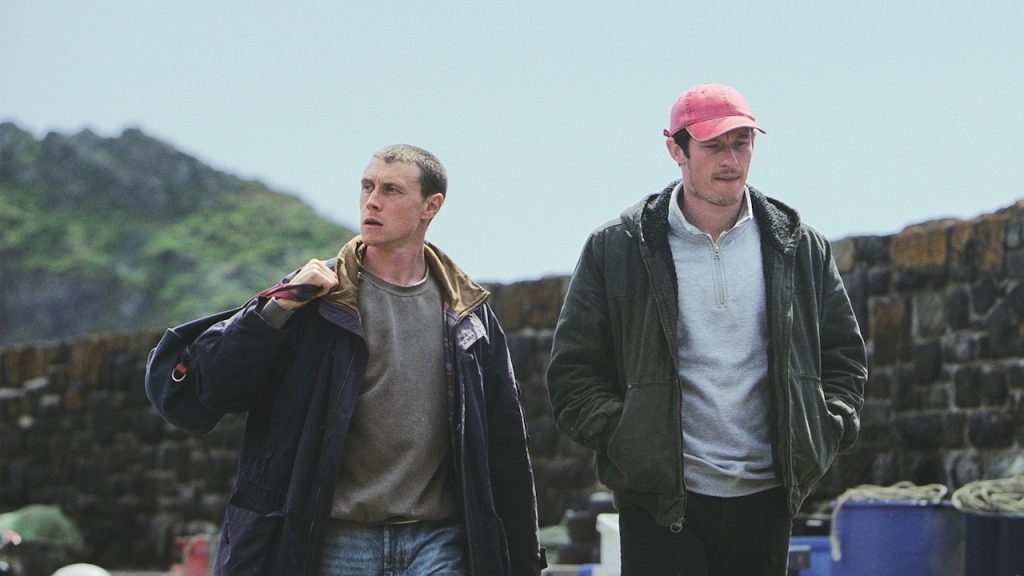Two men walking outdoors near Lynton Cinema, with a stone wall and greenery in the background.