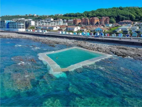 A scenic coastal view of the Little Sea Pool with colourful beach huts and lush green hills in the background.